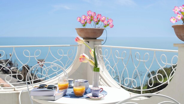 Un balcone con un tavolo apparecchiato per la colazione, un vaso con fiori e vista mare.