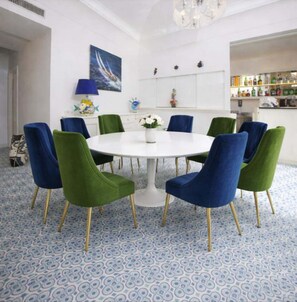 Une table de salle à manger ronde, entourée de huit chaises, un sol blanc orné d’un motif bleu, un lustre et un mur décoré d’un tableau et d’objets décoratifs.