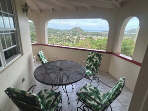 A covered patio with a round table and four chairs, a window with a view of a hilly landscape, and a red trim along the ceiling. - Amazing Antigua Getway (Bolans)
