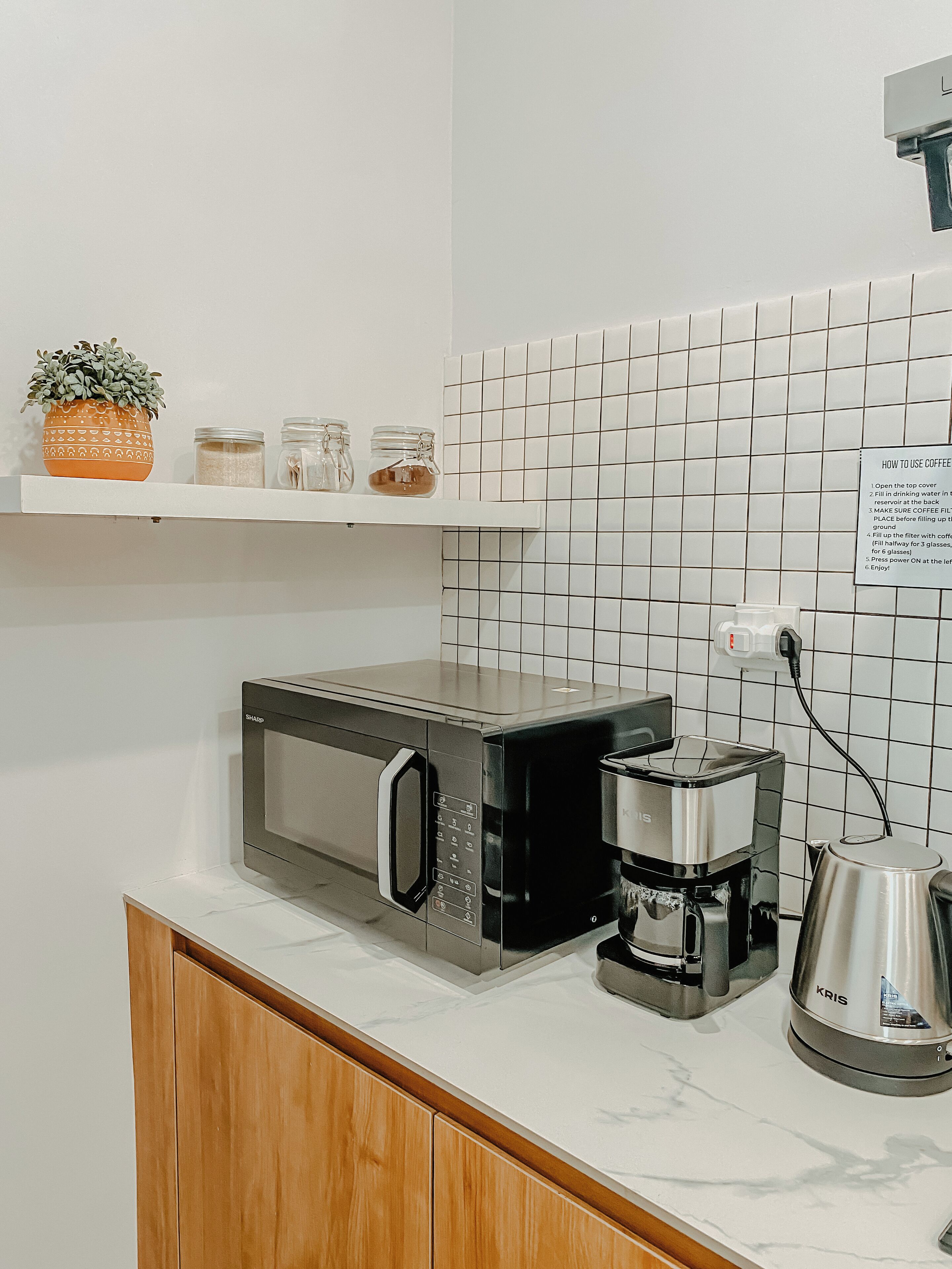 A kitchen with a microwave, coffee maker, and toaster on the counter, a shelf with jars and a plant, and a tiled backsplash.