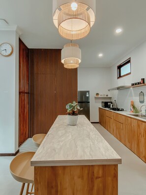 A modern kitchen with a marble countertop, wooden cabinets, a built-in oven, and a hanging light fixture. - The Maru House Seminyak (Seminyak)