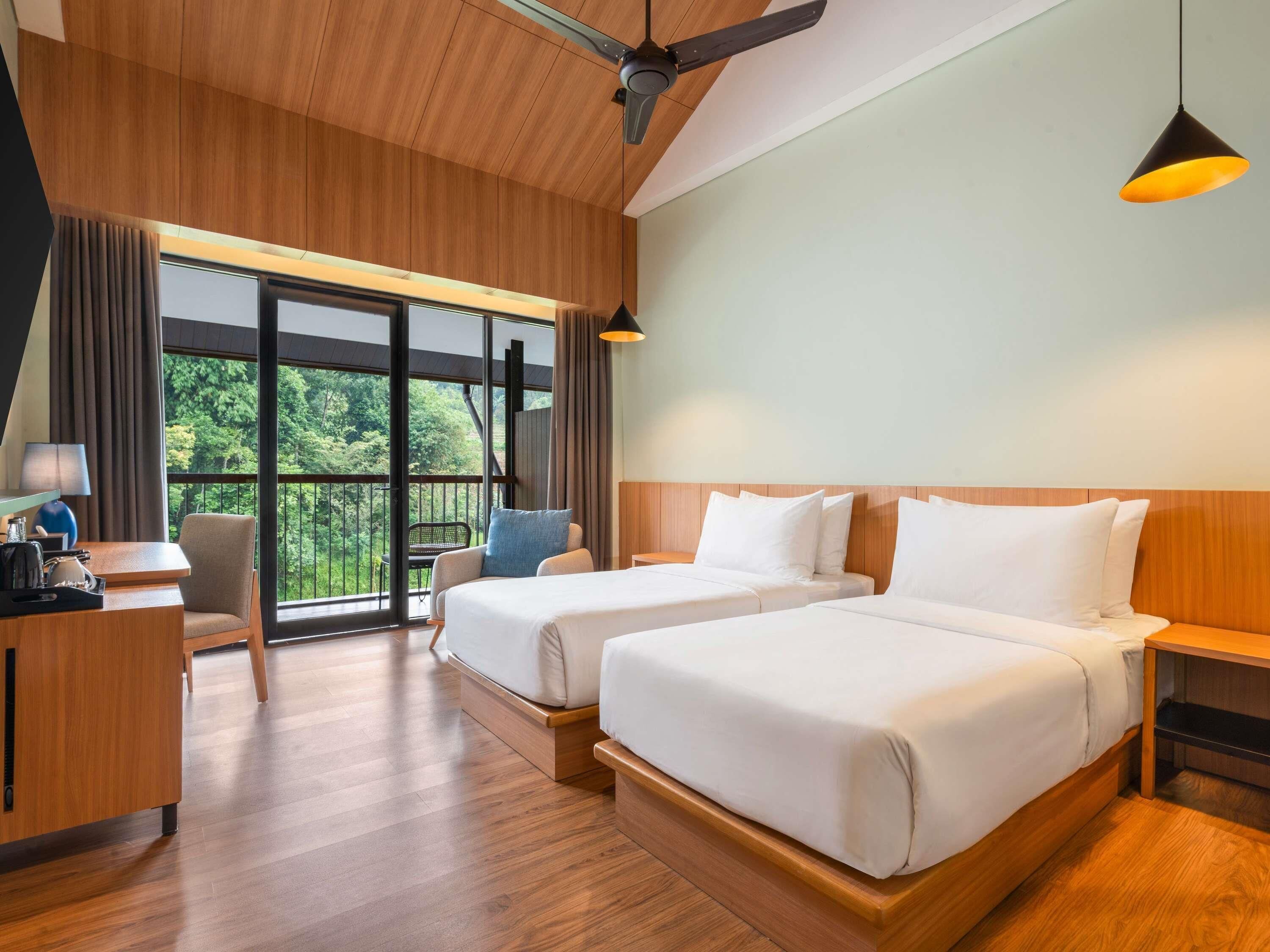 Habitación de hotel con dos camas, un escritorio, una silla y un balcón con vistas a zonas verdes.