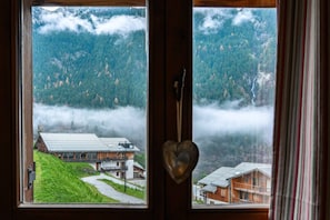 Une vue à travers une fenêtre ornée d’un motif en forme de cœur, donnant sur une région montagneuse avec des bâtiments et un sentier.