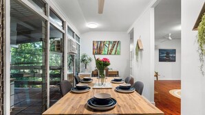 A dining area with a wooden table set for four, surrounded by chairs. There is a vase with flowers on the table, and a painting on the wall. - Zen City Tropics Fenced Yard - Balcony - Desk (Darwin)