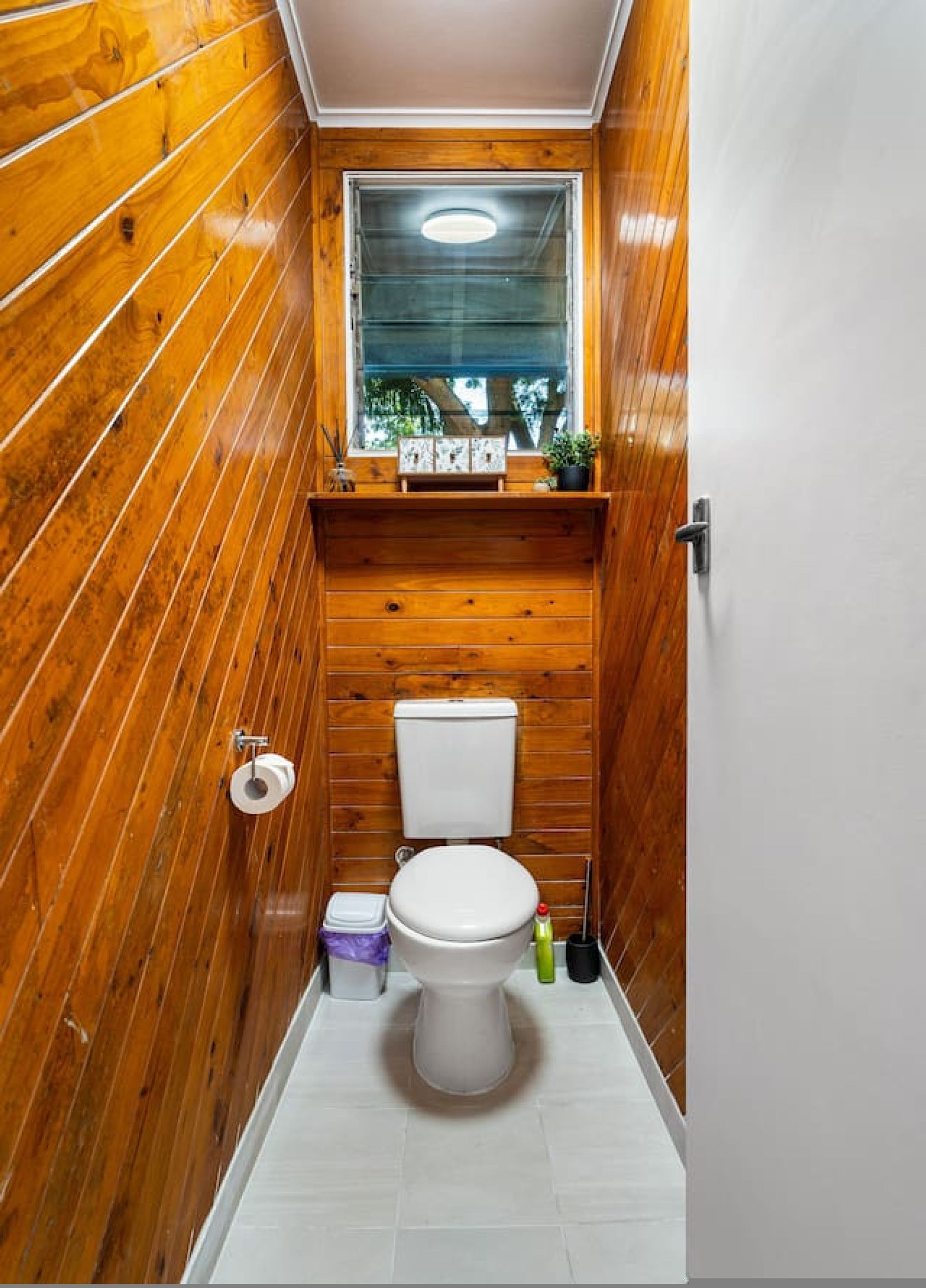 Un baño pequeño con paredes de madera, un inodoro blanco, un ventanal y un estante con artículos de aseo.