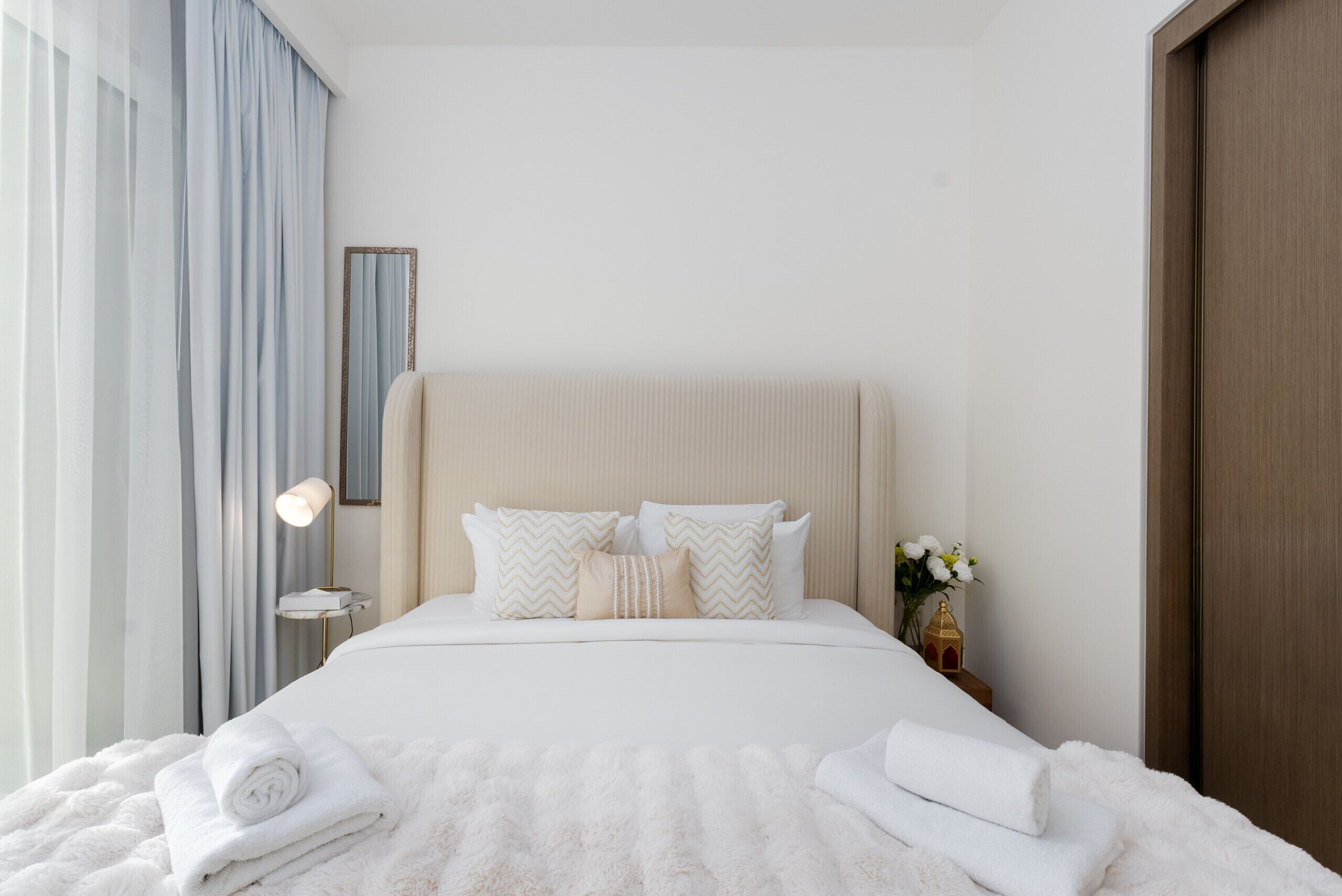 Ein ordentlich bezogenes Bett mit weißen Leinen und einer flauschigen weißen Decke, ein Nachttisch mit Lampe sowie eine Vase mit Blumen.