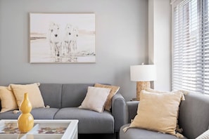 A living room with a grey sofa, beige cushions, a white vase, and a framed picture of horses.