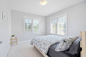 A bedroom with a bed, two windows with blinds, a small wooden side table with a plant, and a ceiling light.