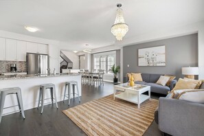 A modern kitchen with a central island, grey sofas, and a chandelier. - Modern House Steps From Dining, Shopping & Lakes (Aurora)