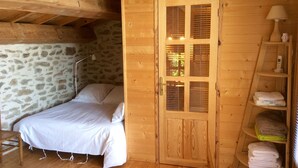 Habitación de cabaña de madera con cama, pared de piedra, puerta de madera con contraventanas y estante con toallas.