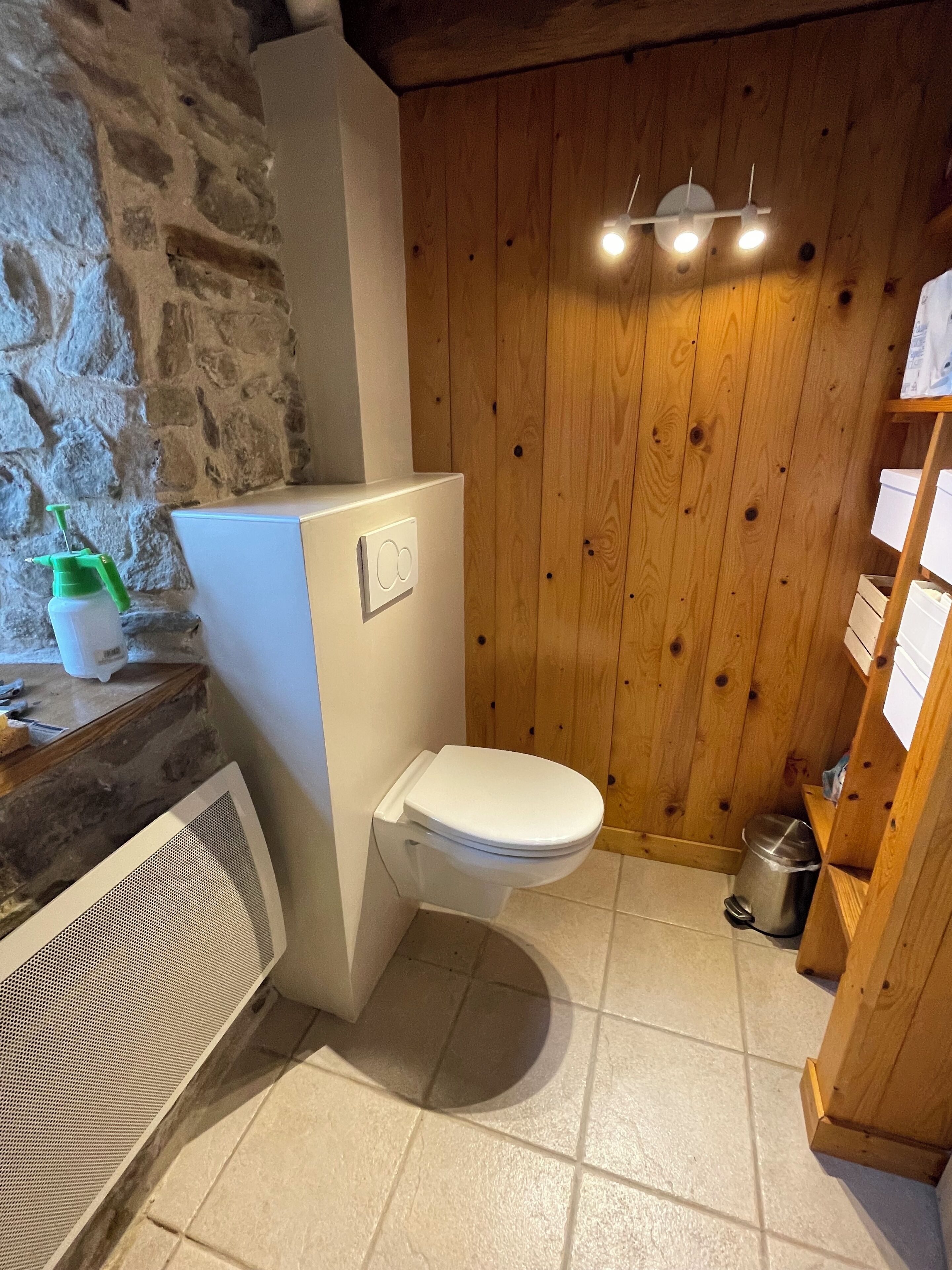 Un baño compacto con inodoro blanco, pared de piedra, paneles de madera y suelo de baldosas.