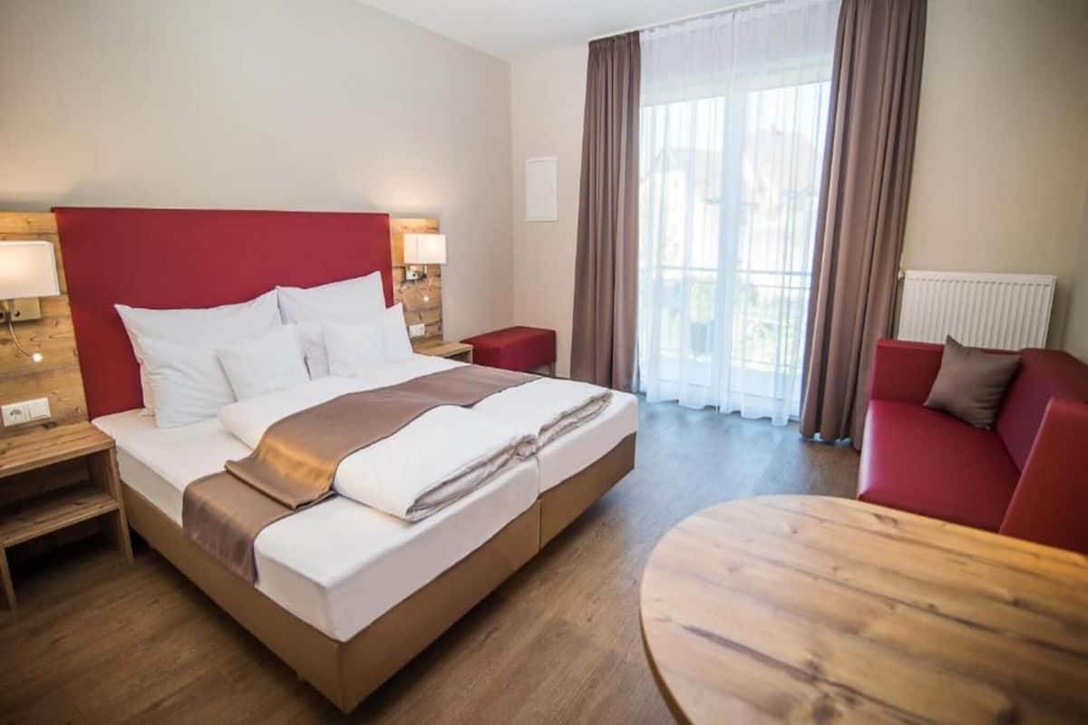 Habitación de hotel con una cama grande, cabecera roja, mesita de noche de madera, sofá rojo, mesa de centro de madera y una ventana con cortinas.