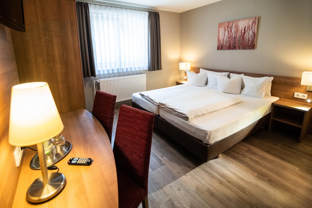 Habitación de hotel con cama, mesitas de noche, un escritorio con lámpara, una silla y una ventana con cortinas.
