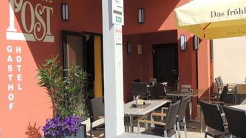 An outdoor seating area with tables and chairs, a sign for 'NEUE POST GHOSTHOFF', and a 'Zimmer frei!' sign indicating available rooms.
