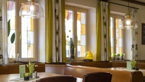 A restaurant with wooden tables and chairs, large windows with green curtains, and hanging pendant lights. - Hotel-Gasthof Neue Post (Leutershausen)