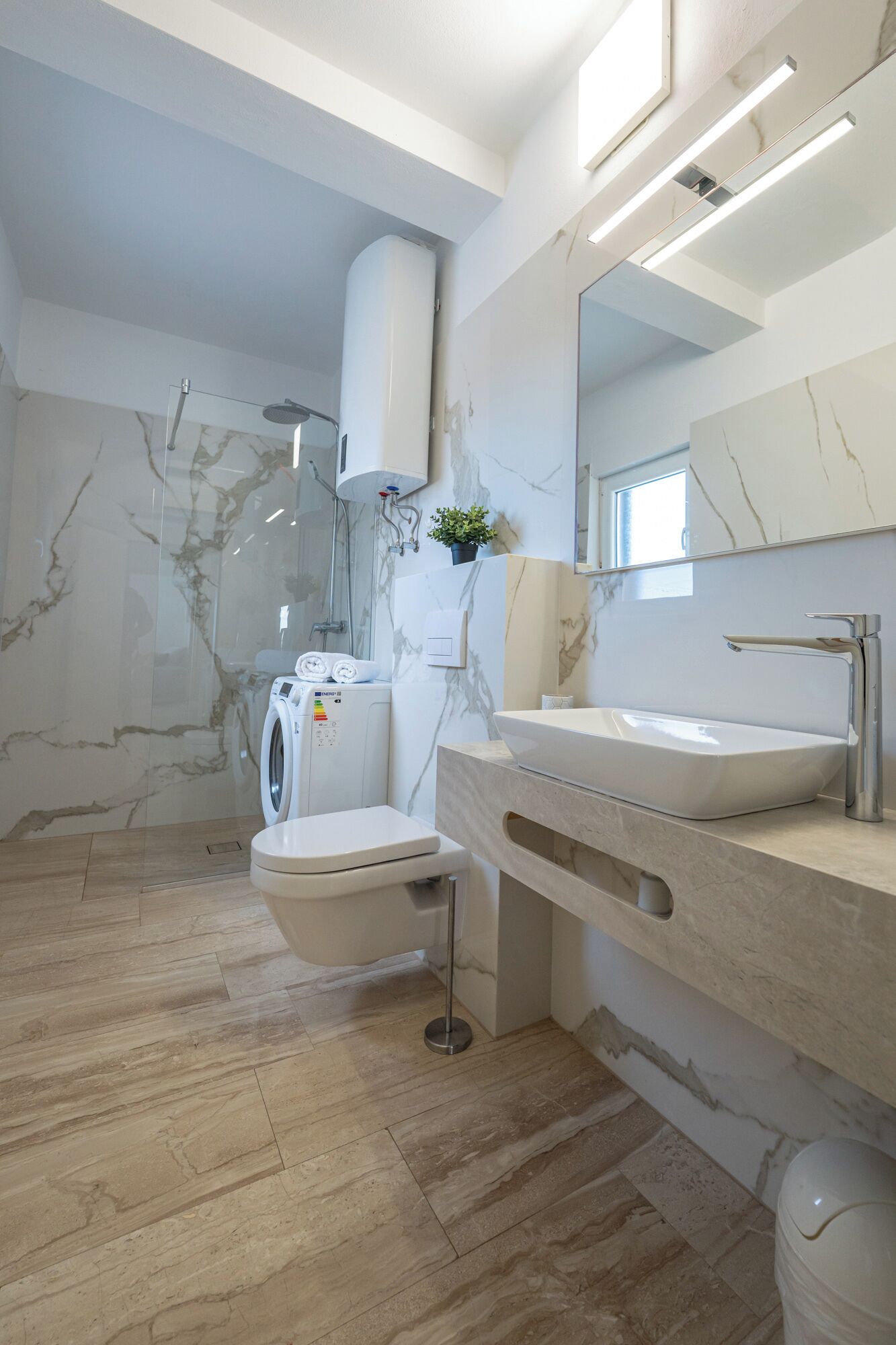 A modern bathroom with a marble wall, a built-in washing machine, a toilet, and a sink with a mirror above it.