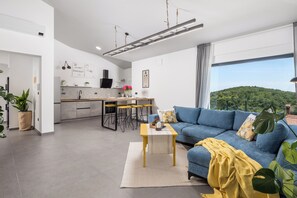 Ein zeitgemäßer Wohnraum mit Küche, Essbereich und großem Fenster mit Blick auf einen bewaldeten Hügel.