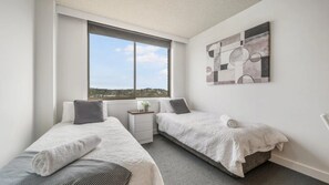 A bedroom with two single beds, a window with a view, a small bedside table, and a wall-mounted abstract art piece. - Fountain Court 24 - Walk to Everything! The Ultimate Coastal Escape in the Heart of It All! (Port Macquarie)