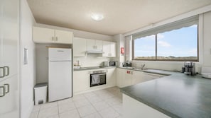 A modern kitchen with white cabinetry, a built-in oven, microwave, and coffee maker, and a window offering a view of the landscape. - Fountain Court 24 - Walk to Everything! The Ultimate Coastal Escape in the Heart of It All! (Port Macquarie)