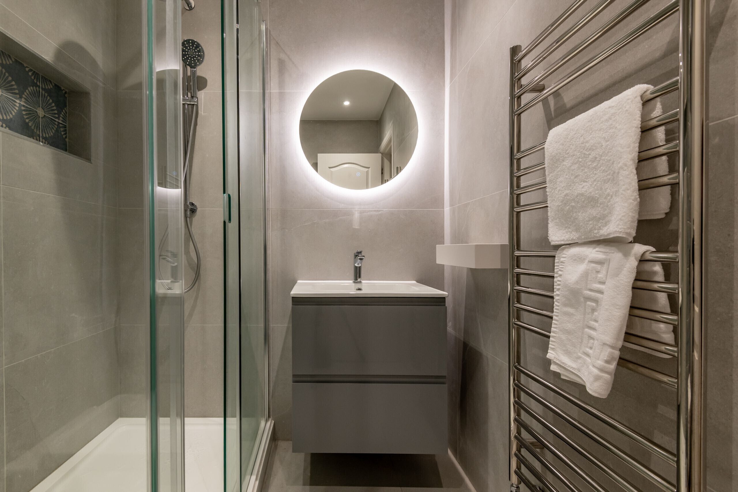 A modern bathroom with a glass-enclosed shower, a round mirror, a sink, and a towel rack with towels.