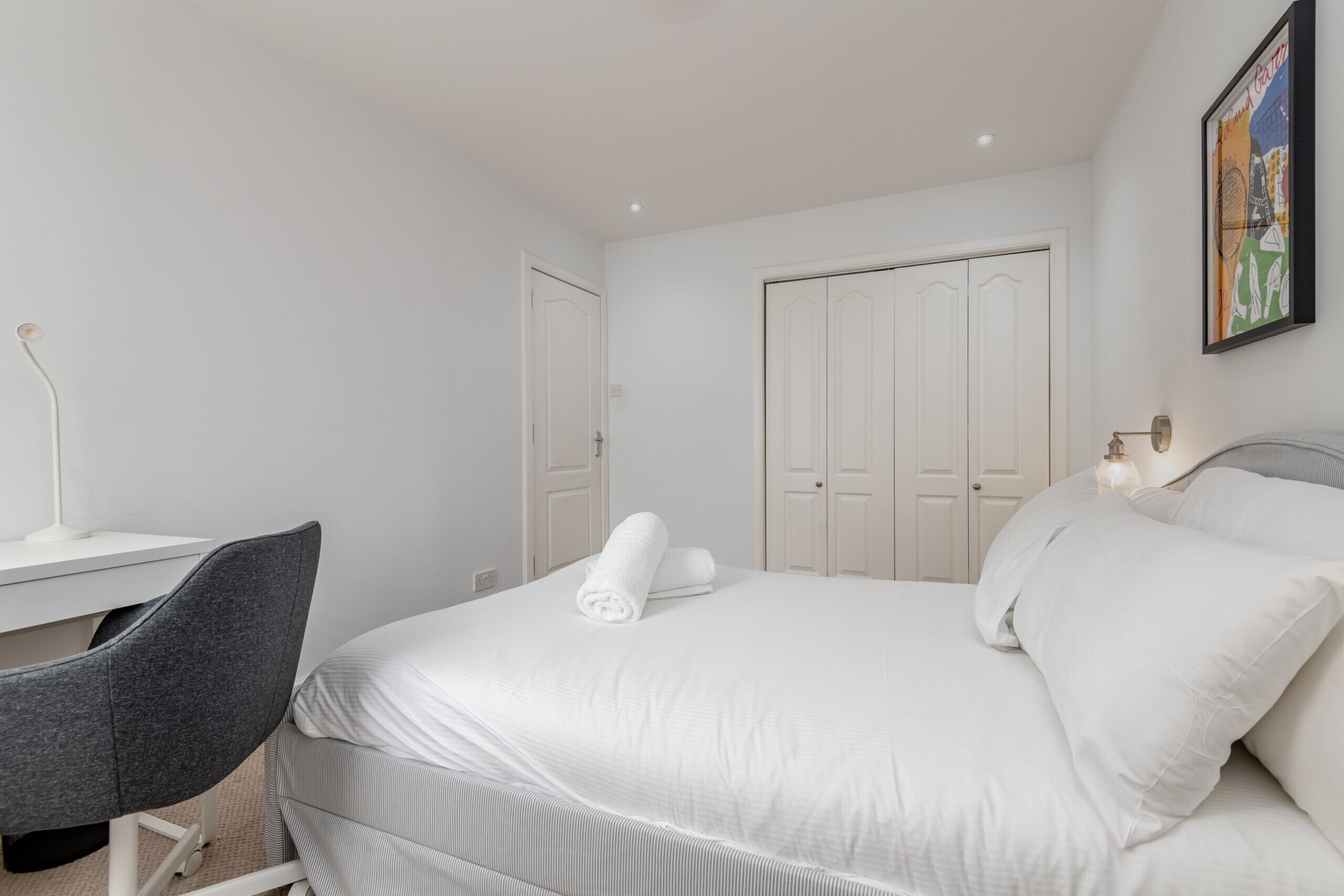 Habitación de hotel con una cama, un escritorio y una silla. Se observan puertas blancas y un cuadro enmarcado en la pared.