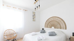 Et soveværelse med en seng, en fletstol, en vægmonteret ventilator og en loftlampe.