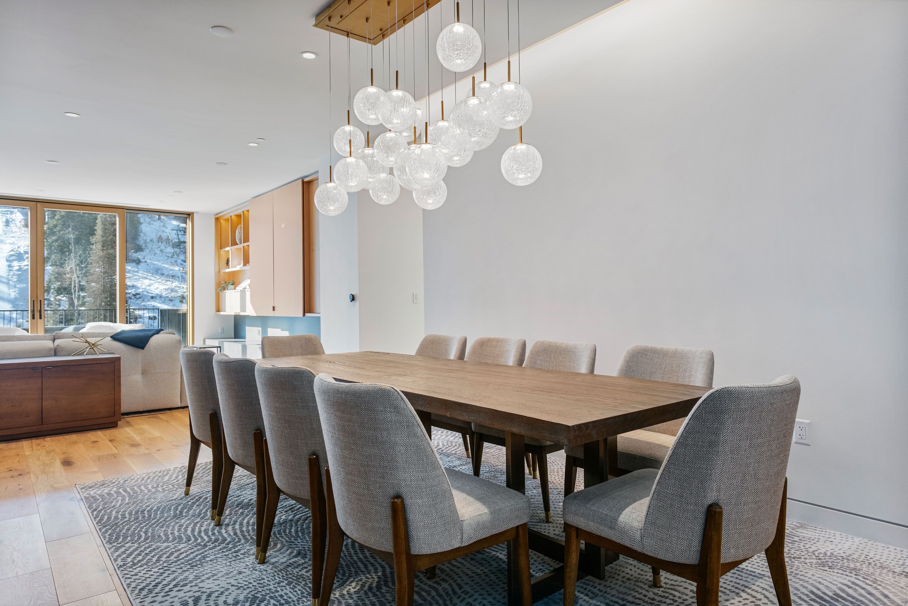Un comedor moderno con una mesa de madera y sillas tapizadas en gris, una lámpara colgante y vistas a un paisaje nevado a través de ventanas amplias.