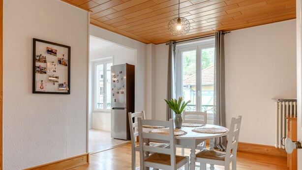 A dining area with a round table, white chairs, a vase with a plant, a bulletin board with photos, and a view of a house through a window. - Apartment Le 47: Spacious Holiday Home with Wi-Fi in Alsace (Thann)