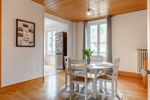 A dining area with a round table, white chairs, a vase with a plant, a bulletin board with photos, and a view of a house through a window. - Apartment Le 47: Spacious Holiday Home with Wi-Fi in Alsace (Thann)