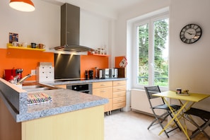 A modern kitchen with a central island, a dining area with a yellow table, and a large window providing a view of greenery.