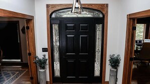 Een zwarte deur met een decoratief glasraam erboven, aan weerszijden omgeven door twee potplanten.