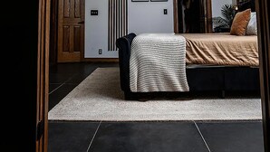 Een slaapkamer met een houten plafond, een bed met een beige deken, een zwarte stoel en een witte vloerkleed.