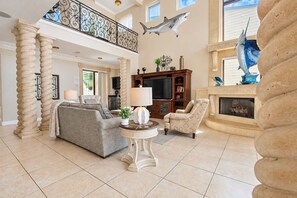 A spacious living room with a high ceiling, a fireplace, and a large window. - Vista del Mar by GoldenCoastRentals | Beachfront Villa • 6BR + Loft • 7 Baths • Game Room (Indian Rocks Beach)