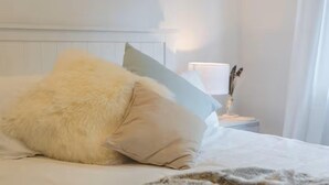 Ein Bett mit einem flauschigen weißen Kissen und einem beigen Kissen, ein Nachttisch mit einer Lampe und dekorativen Federn, sowie ein weißes Kopfstück.