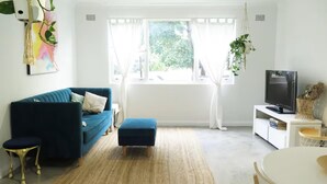 Ein Wohnzimmer mit einem blauen Sofa, einem kleinen Hocker, einem Fernseher auf einem weißen Schrank und einem Fenster mit weißen Vorhängen.