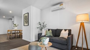 Uma sala de estar moderna com um sofá cinza, uma mesa de centro redonda de madeira, um abajur e uma área de jantar com mesa e cadeiras.