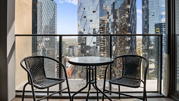 Uma varanda com duas cadeiras de vime preto e uma pequena mesa redonda, com vista para um horizonte urbano com edifícios modernos e altos.