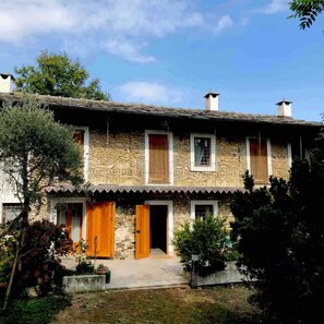 A stone building with two wooden doors and windows, surrounded by greenery. - Mombracco Retreat | Stone & Woods | Saluzzo (Envie Piemonte)