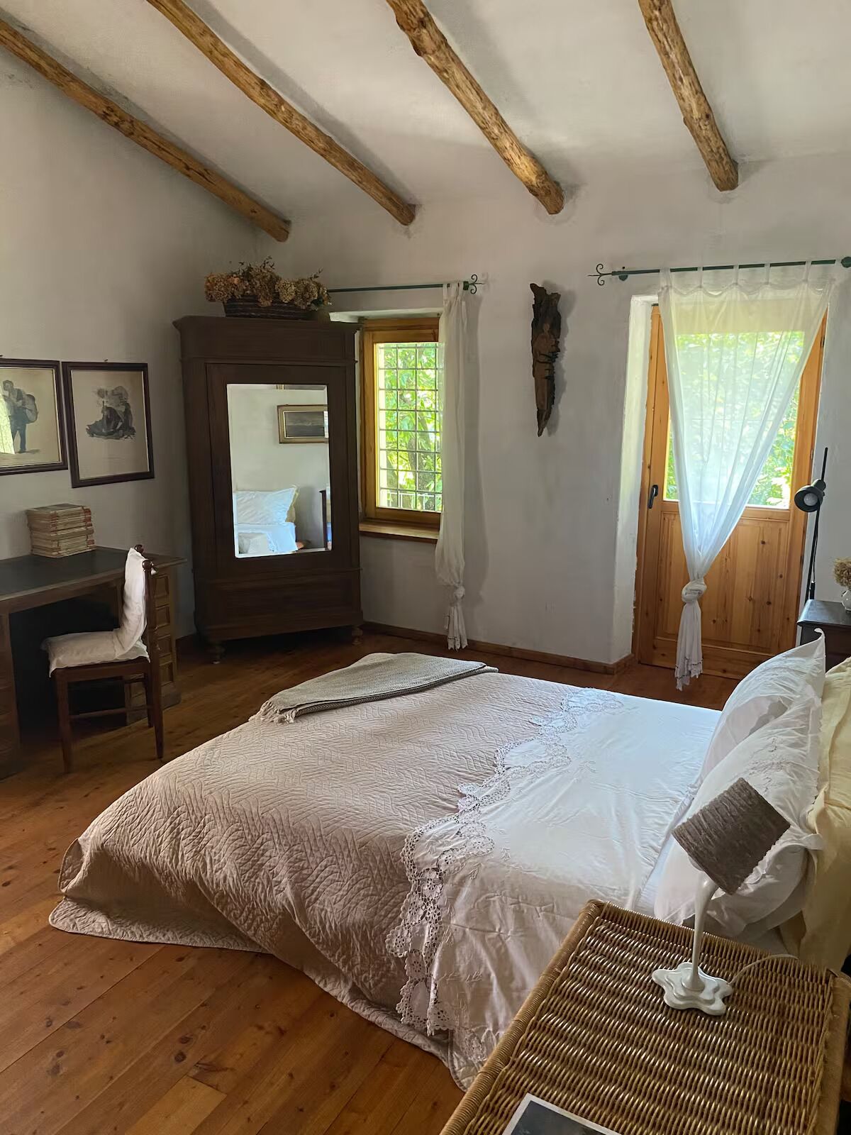 Un dormitorio con una cama grande, vigas de madera, un escritorio con silla, una ventana con cortinas y cuadros enmarcados en la pared.