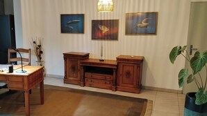 Un salon avec un bureau en bois, une chaise, un canapé et une grande plante en pot. Trois tableaux encadrés sont accrochés au mur.