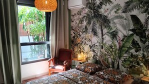 Une pièce comprenant un lit, une chaise, une fenêtre avec des rideaux et un mur orné d’une fresque aux motifs tropicaux.