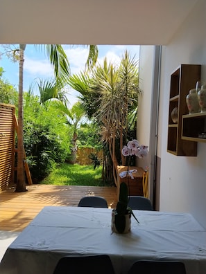 A room with a table covered by a white tablecloth, a potted plant, and a view of a lush garden with palm trees and a bamboo fence. - Vacation apartment with terrace and small garden (St. Leu)