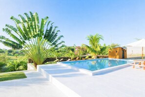 Une piscine à l’eau d’un bleu cristallin, entourée de carreaux blancs, de palmiers et d’un petit bâtiment.