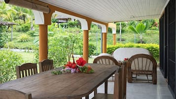 An outdoor dining area with a wooden table and chairs, surrounded by lush greenery and a thatched-roof structure.