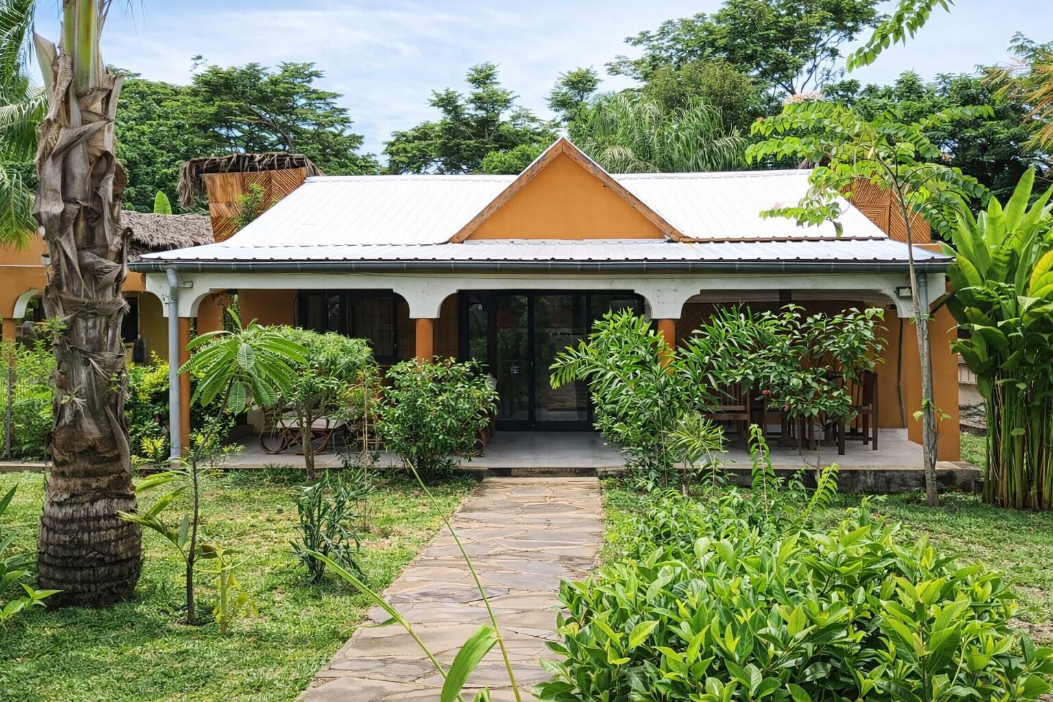 A two-story building with a covered patio area, surrounded by lush greenery and a stone pathway leading to the entrance.