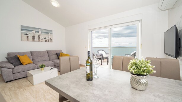 A modern living room with a grey sofa, a white coffee table, and a dining table. There is a painting on the wall, a bottle of wine, and a potted plant on the table. - Stylish Two-Bedroom Apartment with Spacious Sea-View Terrace & Shared Pool (Novalja)