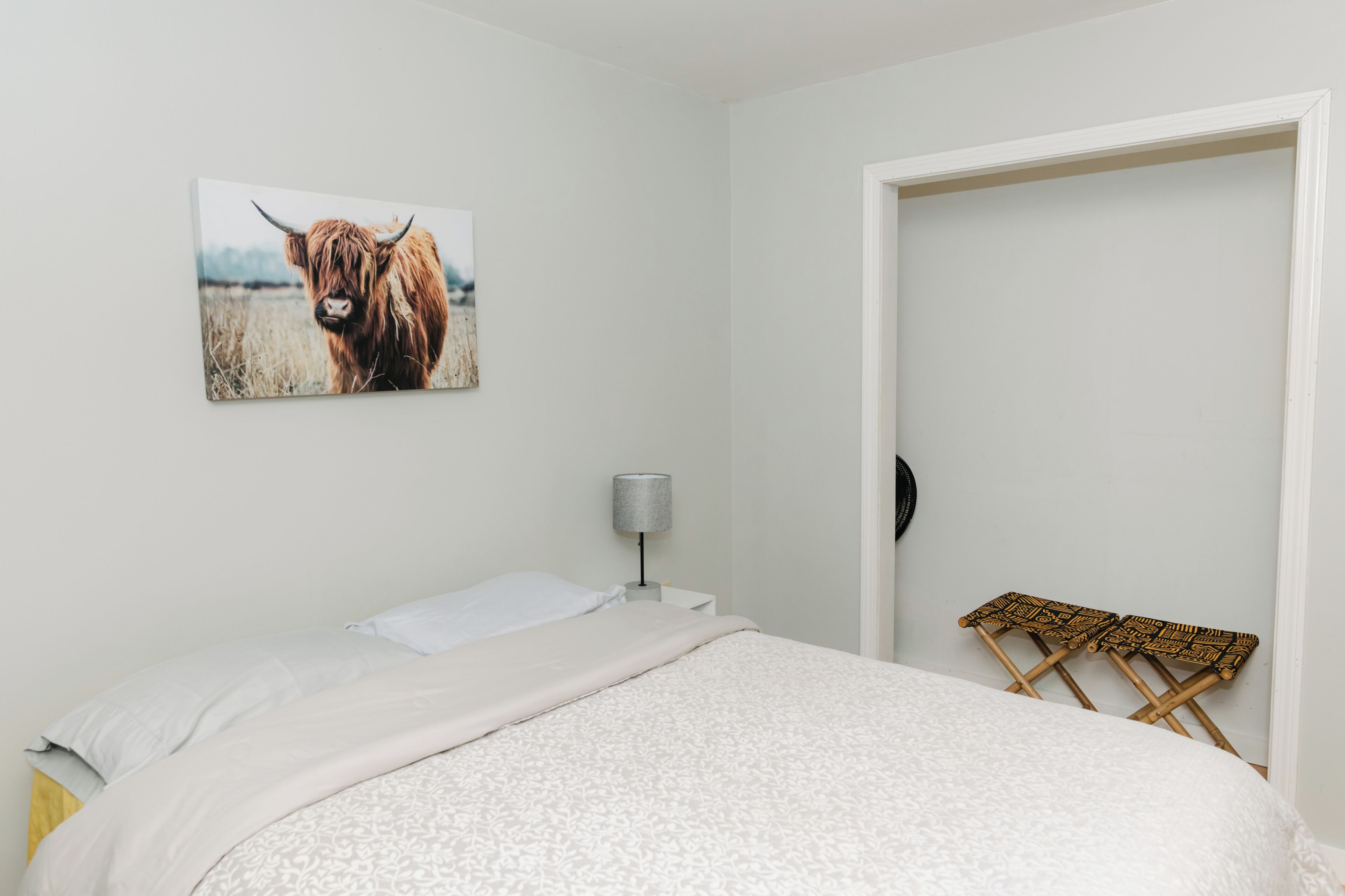 Une chambre à coucher avec un lit, une lampe, une petite chaise et un tableau représentant une vache accroché au mur.