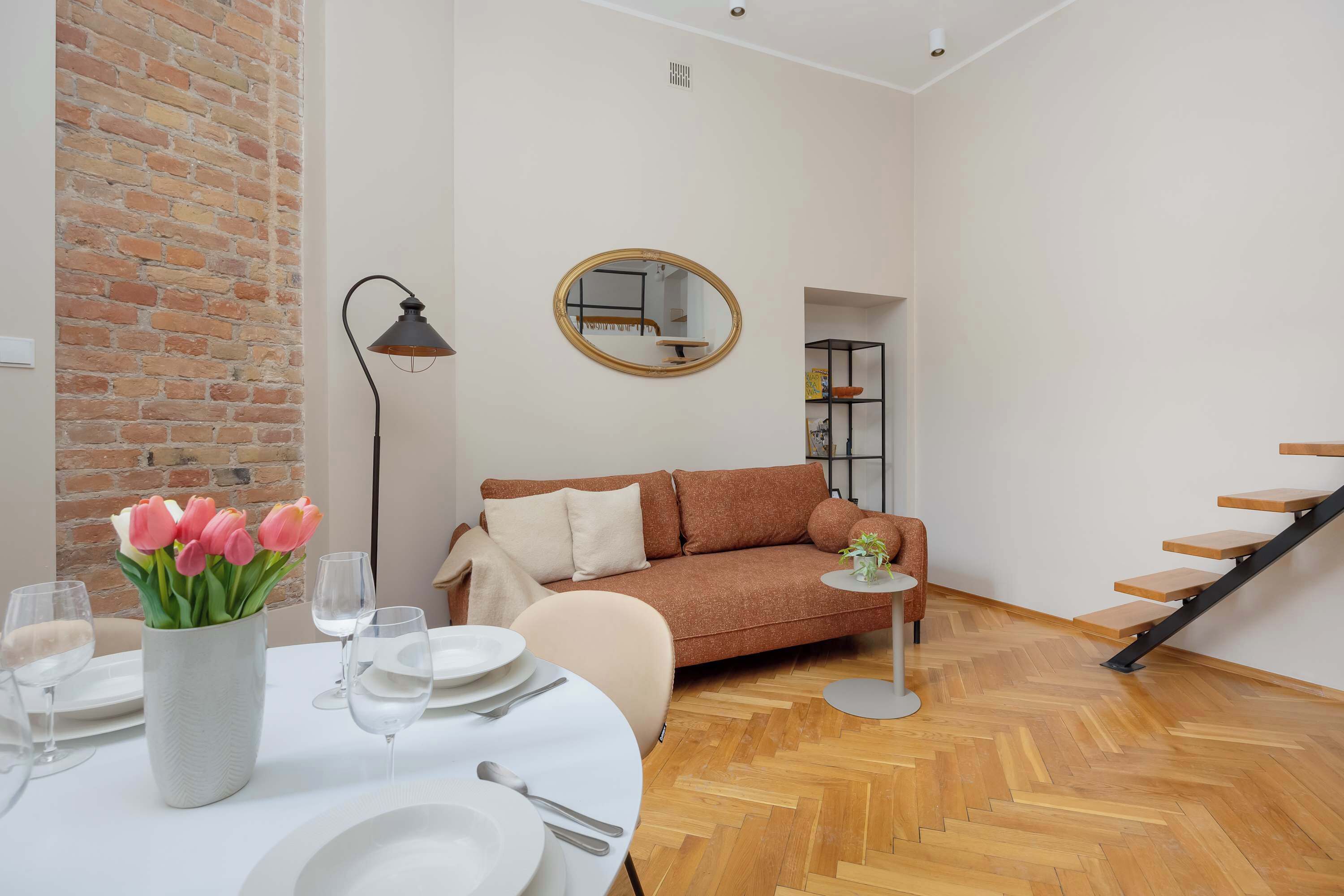 A modern living room with a brick wall, a round mirror, a sofa, a dining table set with plates and glasses, and a staircase.