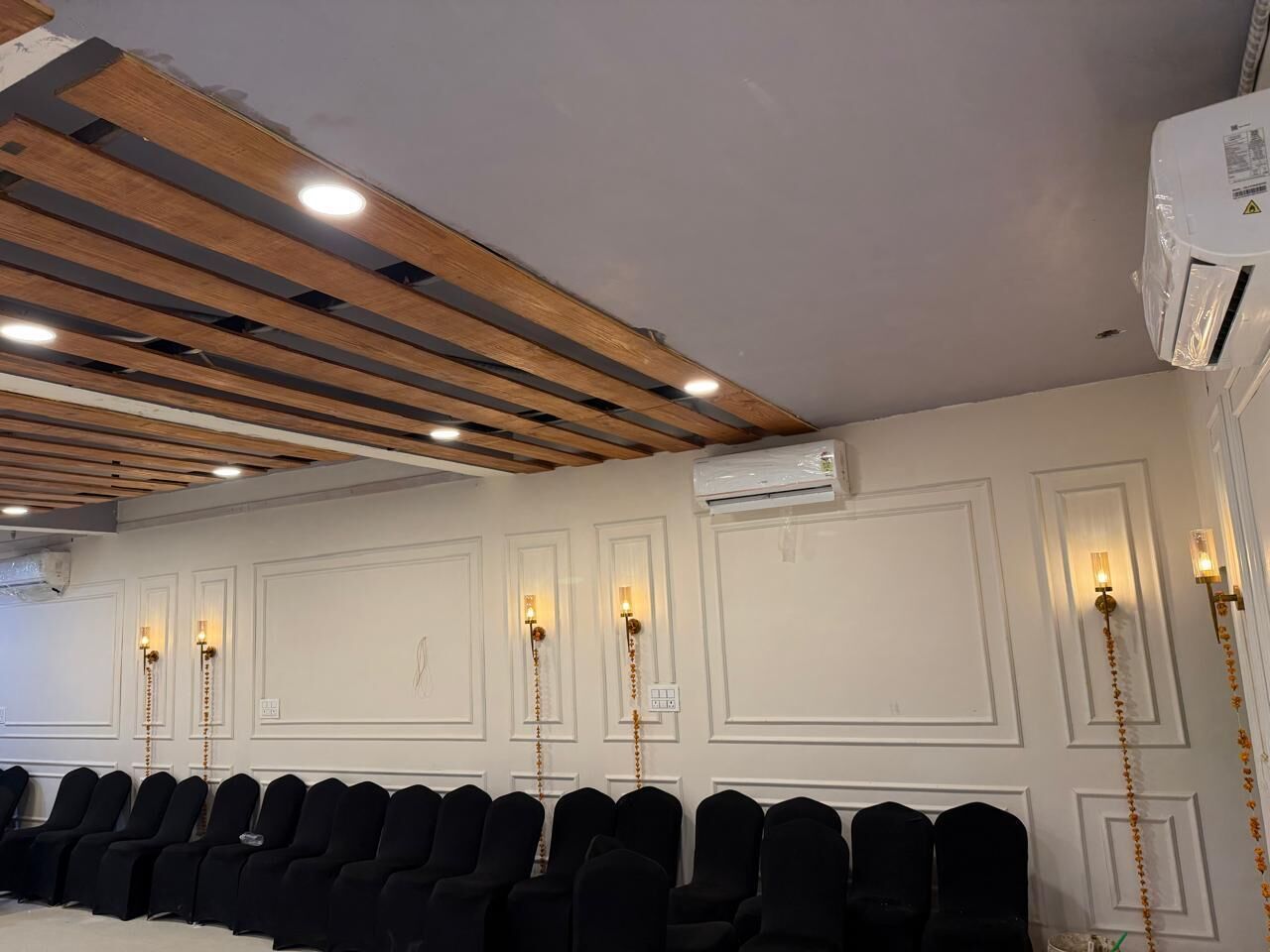 A conference room with rows of black chairs, wooden ceiling beams, white walls with paneling, and wall-mounted lights.