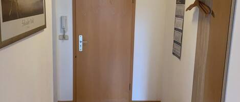 A hallway with a wooden door, a framed picture on the wall, a mirror, and a small mat on the floor.